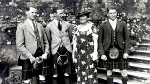 Black and white photo of three men in kilts, either side of a lady - their mother - in a floral dress.