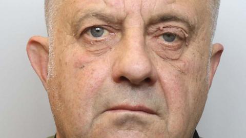 Police mugshot of Trevor Leach. He has close-cropped grey hair and grey stubble. Standing in front of a grey backdrop, he stares at the camera with an impassive facial expression.