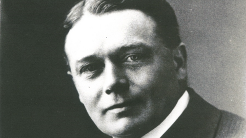 A black and white photo of a man in a suit and shirt. He has short dark hair, swept back and centre-parted and is clean shaven.