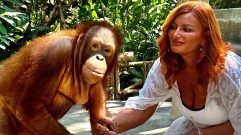 Kelly Kershaw is kneeling down in a white dress holding the hand of an orangutan, which is looking at the camera. Behind them is a forest environment.