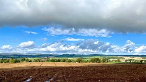 large band of cloud sits across the whole of the scene with ah layer of blue sky and clouds below, with mountains, trees and muddy brown fields on the land.