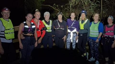 The image shows a group of women standing together at night. They are all smiling at the camera, wearing hi-vi clothing.