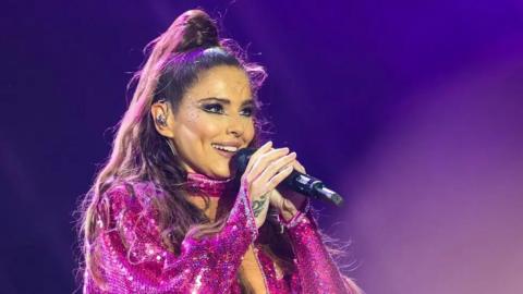 Cheryl singing into a microphone with a purple background. She wears a sequined pink costume and has sequins stuck on her cheek. There is an earpiece in her ear and her hair is half up in a knot on top of her head with the rest cascading down past her shoulders