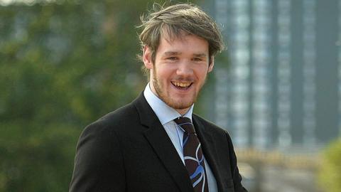 A smiling young man who has sandy hair and a beard, wearing a black suit jacket, a light blue shirt and a brown and blue patterned tie. He is outdoors, with trees and a tall building blurred in the background.