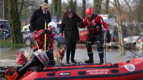 Rescue personnel in red clothing assist a resident in a brown coat onto a red inflatable boat