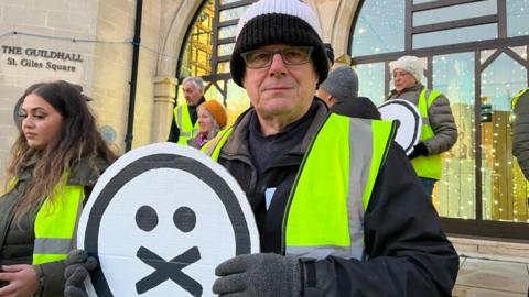 Man wearing woolly hat and yellow hi-viz holds a silent protest sign