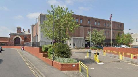 A Google street view screenshot showing the entrance into HMP Bristol prison. It is a large boxy looking building with inset windows. There are yellow railings and an electric barrier arm outside. There is a red brick wall that surrounds the perimeter of the prison and a couple of trees and bushes growing around the car park.