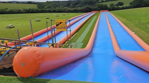 The image shows the top of four slip-and-slides situated on a green field in a rural area. They are blue plastic strips spreading out ahead and going downhill, with inflatable orange tubes on either side.