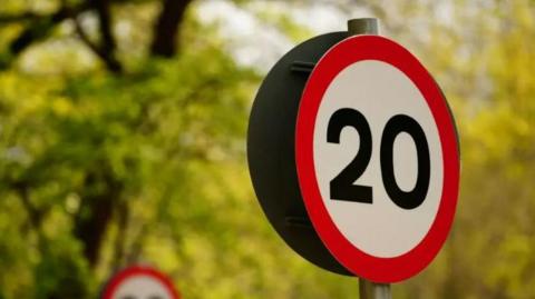 A red and white road sign with 20 written on it in black. Behind the sign are trees