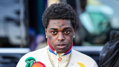 Kodak Black stares directly into the camera. He is wearing a white knitted quarter zip with a high neck that has blue and pink stripes on the collar.