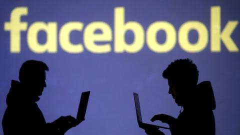 Silhouettes of laptop users are seen next to a screen projection of the Facebook logo.