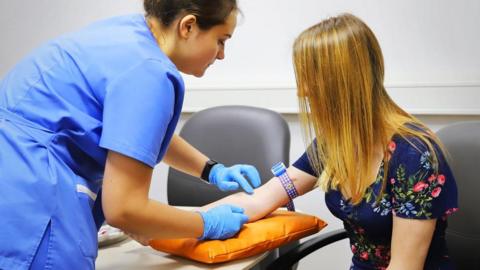 A female nurse with short dark hair, wearing a light blue uniform, leans over a female patient while wearing blue latex gloves which she is wearing on her hands as she prods the woman's right arm, which is resting on an orange cushion. The woman she is prodding has long light brown hair, which hangs over her face, and wears a dark blue top with a pattern of flowers on it.