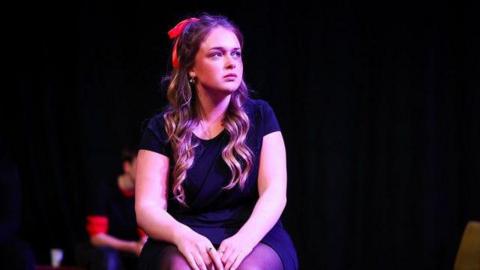 A young girl with curly hair and wearing a black t-shirt and a black skirt sitting on a chair and looking at something out of shot.