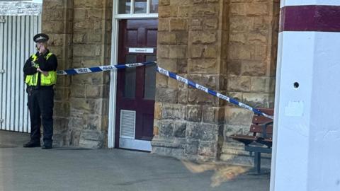 A police officer stands outside a gents' toilets, which has police tape across the door.
