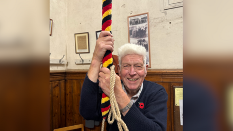 Jack is smiling to camera in the bell tower, holding onto a rope which is black, red and yellow-striped. He has white hair and is wearing a blue jumper with a poppy attached. There is a black and white photo behind him showing old pictures of bell-ringers.