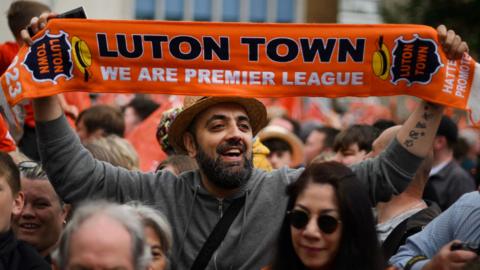 An Asian man is among of crowd of Luton Town fans and he is holding up an orange scarf above his head that says "Luton Town, we are Premier league" on it. He has a dark beard and is wearing a straw hat and a grey hooded zip up jacket.