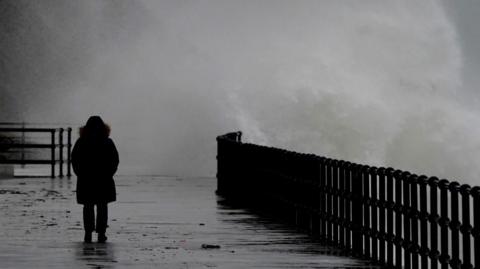 A silhouetted person walks along a paved seafront wearing a raincoat. To the right of the image, sea splash from a large wave spills over a silhouetted fence on to the pavement behind the person. Conditions appear wet, windy and grey.