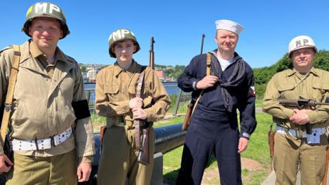 Four men are standing dressed in World War 2 style clothing. Three of the men are wearing green army uniform while the other is wearing a navy sailors outfit, they are all wearing a head piece. Three of the men are holding visible weapons. Behind the men is an area of grass and a section of the river Foyle.  