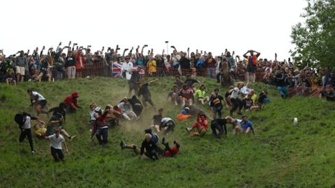 A large crowd of people falling down a steep grassy slope chasing a wheel of cheese