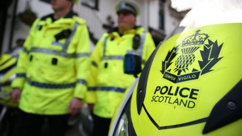 The front of yellow motorbike emblazoned with the Police Scotland logo. In the background are two police officers wearing fluorescent yellow police jackets.