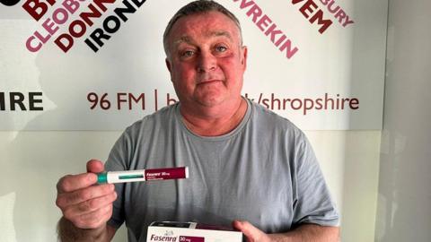 A headshot of man in a grey t-shirt, holding a red and white dispenser that is a new drug to treat asthma. He is also holding a medicine box that reads "Fasenra" in burgundy writing. 