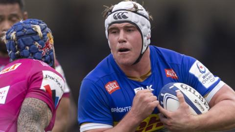 Ernst van Rhyn, with ball tucked under his arm, braces himself to barge past an Exeter Chiefs covering defender who is stooping to make a tackle