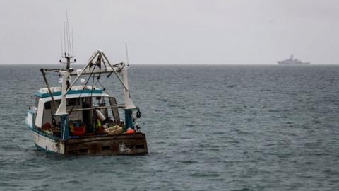 A fishing boat off the coast of Jersey with HMS Tamar in the background on the horizon