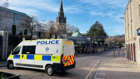 A police van parked across a road, with shops in the background.