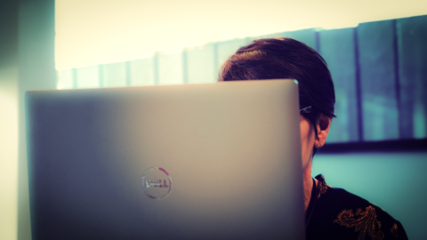 An anonymous lady sits behind a computer laptop screen, you can only see her short dark hair over the top of the laptop