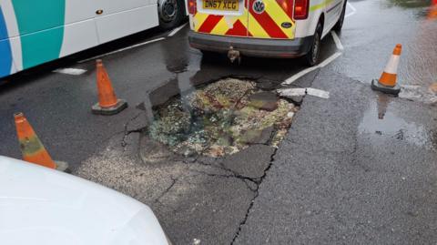 Part of a road surface is badly damaged and there are cones around it. There is a lot of water on the road and a utility company van is parked near the damaged surface.