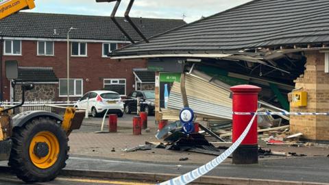 A police cordon around a small village shop which has been rammed into. There is a large hole in the entrance where an ATM would be and the shutters have been torn down.