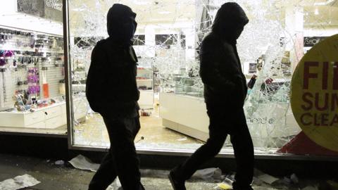 Two men with their faces covered walk past a looted shop during the riots in 2011