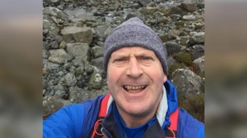 Neil McGibbon taking a selfie, wearing a grey hat on a rocky landscape