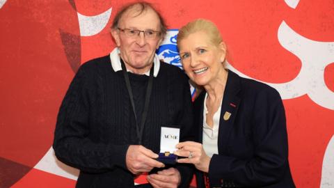 Howard Bevan being presented with a golden whistle by Debbie Hewitt while standing in front of a large England FA badge