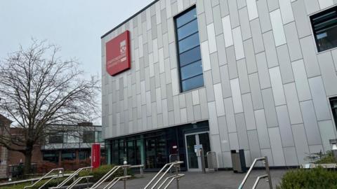The front entrance of a large grey university building, with a red sign on it that says "Staffordshire University".