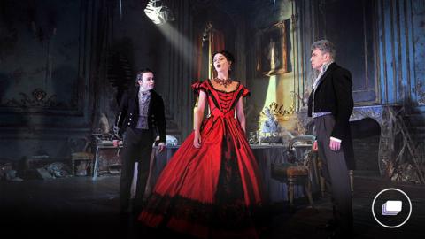 A photograph from the Great Expectations play in London. A woman is in a red and black black gothic ballgown, in between two males in suits. They are in a gothic house. 