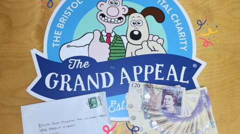 An envelope and several £20 notes sit on top of the logo for Wallace and Gromit's Grand Appeal. The logo is blue and has drawing of Wallace and Gromit.