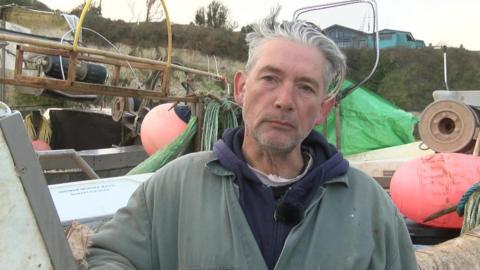 Fisherman Stephen Andrews wearing a faded green overall looks at the camera. He is standing in front of a fishing boat.