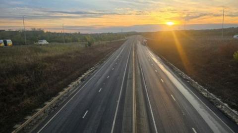 A picture of the A421 carriageway. The photo is taken from a bridge over the carriageway. The road is clear and traffic can be seen heading onto the road via a slip road. The sun can be seen rising in the distance. 