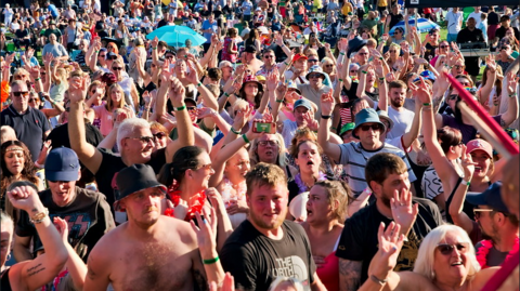 The images shows a festival crowd in the sunshine. There are men and women, some with their hands in the air, men with no tops on and other in sun hats or caps on and a blue umbrella in the background.