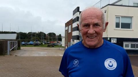 Phil Pollard smiling for the camera on the street on a cloudy day. He is wearing a dark blue shirt branded "Portsmouth Joggers"