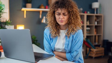 Woman with curly brown hair sits at her laptop clutching her stomach in pain. She has closed her eyes and is grimacing.