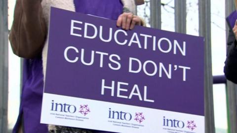 An striking INTO member holds a placard that reads: EDUCATION CUTS DON'T HEAL