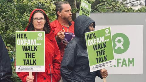 Three protesters with placards next to the Oxfam sign at the charity's headquarters. They are wearing rain coats.
