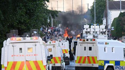 Police landrovers pictured as protests take place. There are several police landrovers, all white with police markings in yellow, orange and blue. In the background, there are lots of people and flames and smoke.