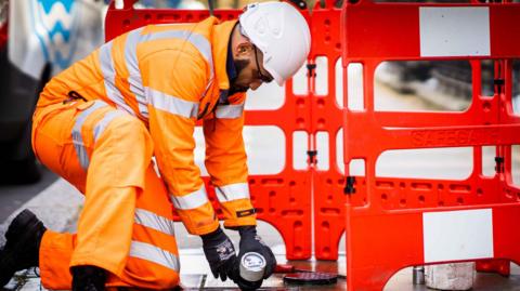 Thames Water employee installing a smart meter on a street. He is wearing an orange high vis outfit and a helmet. There are barriers around the spot where the meter is being installed.