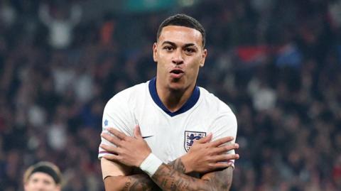 England's Morgan Rogers celebrates scoring for England. He has tightly cropped black hair and has his tattooed arms folded across his chest, which wears a white top with the England logo on