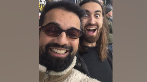 Raul Kohli smiling into the camera in the football stands with brown sunglasses on. He has dark hair and a dark beard and is wearing a beige fleece. On his right is Thomas de Bernede, a man with long brown hair and a beard, smiling with a wide mouth and wide eyes.