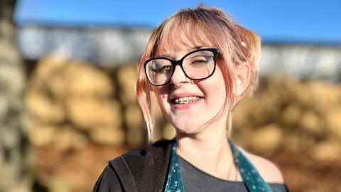 Head and shoulders photograph of Lee taken outside in the sunshine. Lee has blonde hair with pink highlights and is wearing it in a pony tail. She has black glasses on and braces on her teeth. She's smiling. The background is blurred.