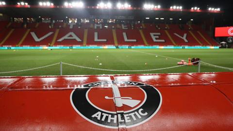 A view of Charlton Athletic's empty football stadium, The Valley, at night under bright floodlights. In the foreground, the top of a red dug-out seat or tunnel structure is prominently featured, displaying the circular Charlton Athletic crest with a hand holding a sword. Beyond the green pitch, red stadium seating spells out "The Valley" in large white lettering across the main stand.